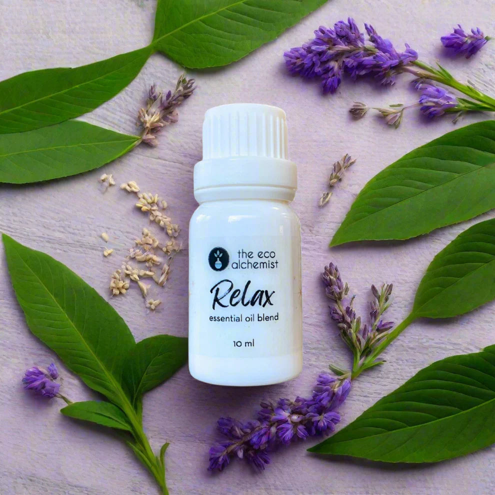 A small white bottle labeled 'Relax essential oil blend' from The Eco Alchemist, surrounded by green leaves and purple flowers.